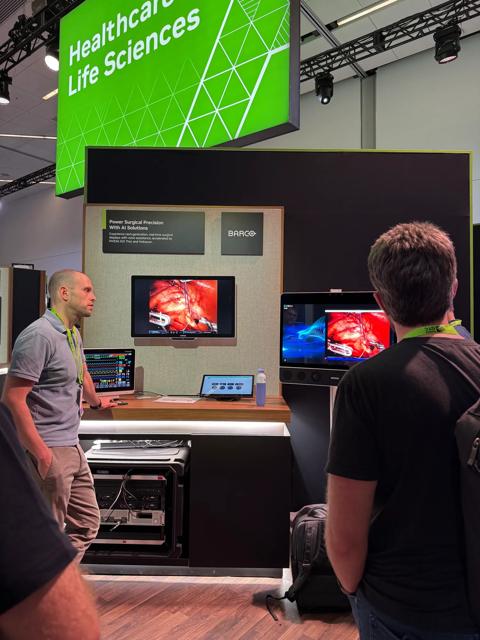 Barco demonstration photograph showing a portrait-oriented surgical display and two participants at the exhibition booth.