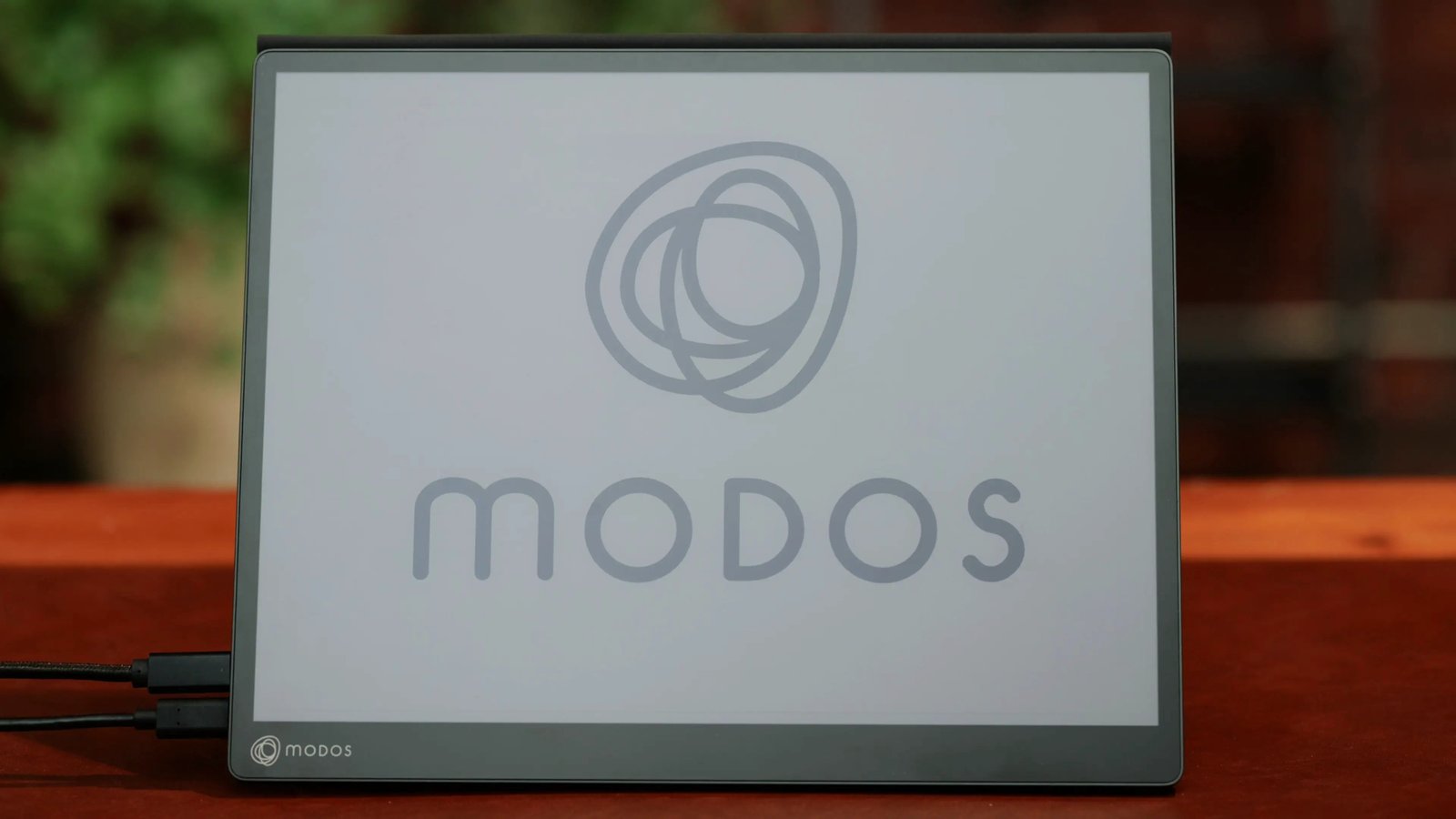 Modos Flow photographed on a wooden table with the Modos logo displayed on its e-paper screen.